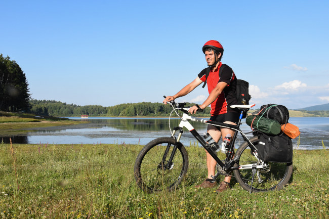 Mann steht mit seinem bepackten Fahrrad vor einem See